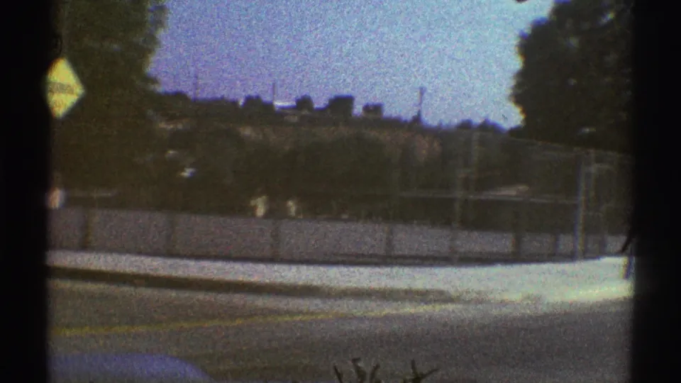 Late-1960s Woodland Hills suburban driving view with a fenced roadside, tree line, utility poles, bright sky, and low hills.