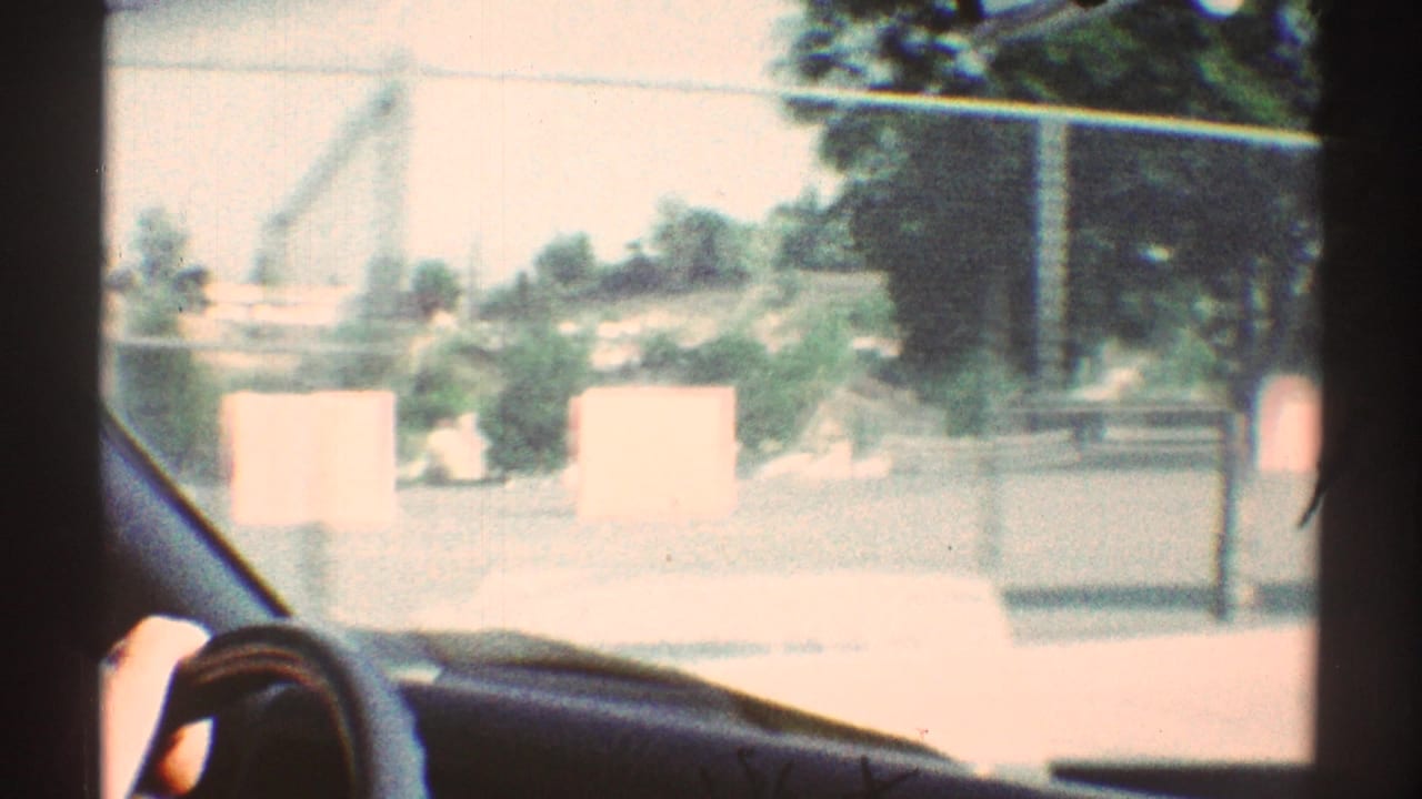 1969 Woodland Hills car-window view passing a fenced school or playground area, with a hand near the steering wheel and sunlit pavement outside.