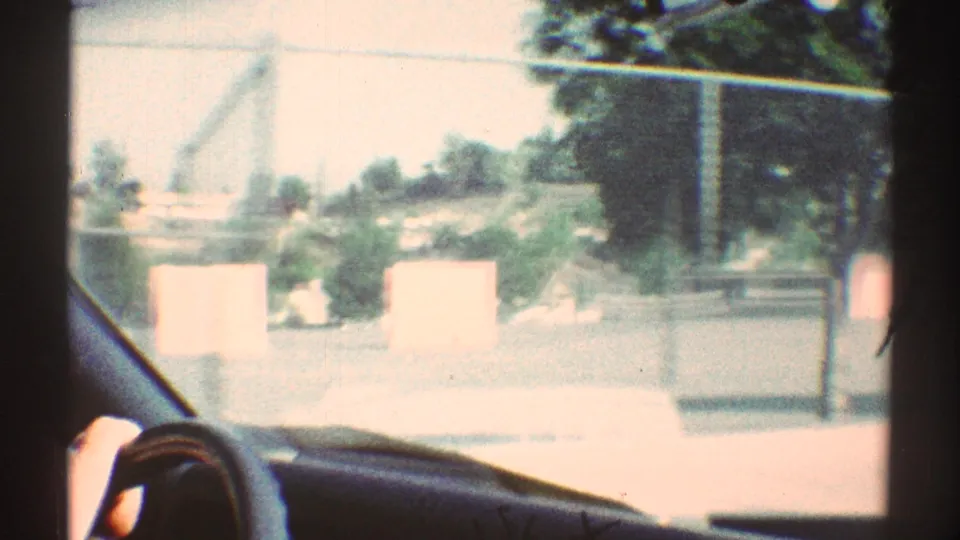 1969 Woodland Hills car-window view passing a fenced school or playground area, with a hand near the steering wheel and sunlit pavement outside.