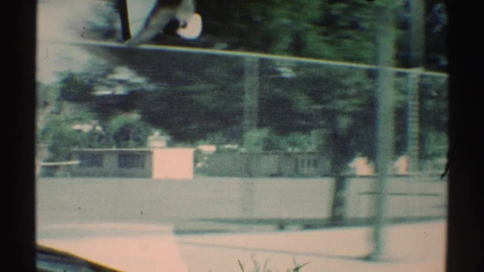 Motion-blurred 1969 Woodland Hills view past a fenced school or playground area, with vertical poles, trees, low walls, and open pavement.