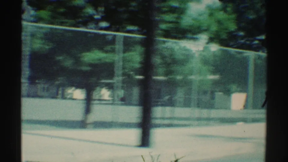 Late-frame 1969 Woodland Hills view from a moving car past an empty fenced schoolyard or playground with trees, a utility pole, and low wall.