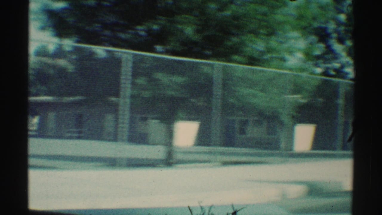 View from a moving car past a chain-link fence, trees, pavement, and low buildings.