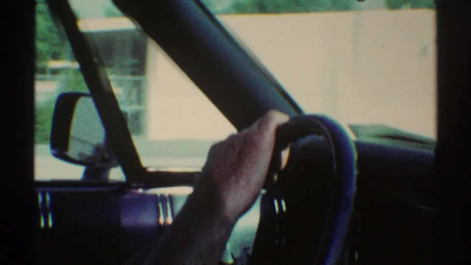 1969 Woodland Hills car interior with a driver’s hand gripping the steering wheel as fencing, pavement, and greenery pass outside.