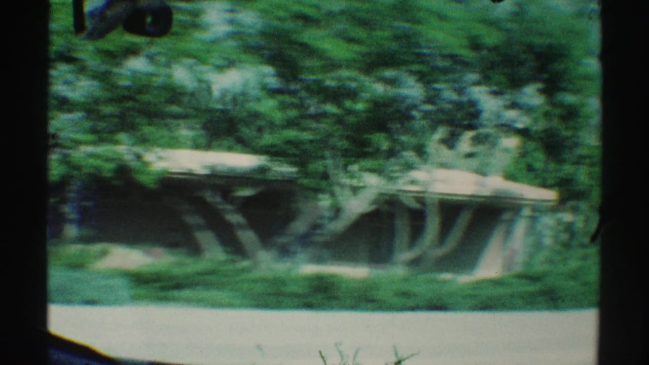 Blurred car-window view of trees, lawn, sidewalk, and a low structure passing outside.