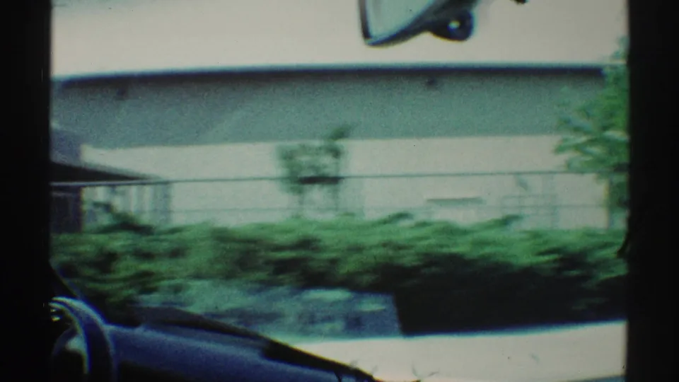 1969 Woodland Hills roadside view with a pale low building, hedges, sidewalk, and part of the car interior in motion blur.