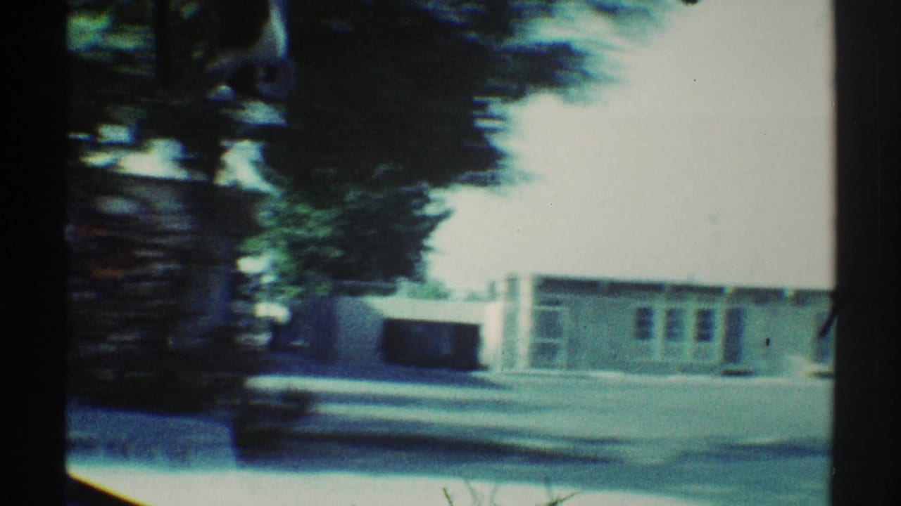 View through a car windshield of trees, pavement, and low school buildings across the street.