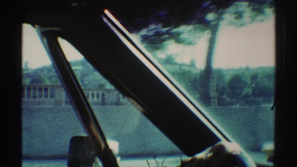 1969 Woodland Hills car-window frame showing windshield supports, a hand near the steering wheel, trees, a low building, and distant hills.