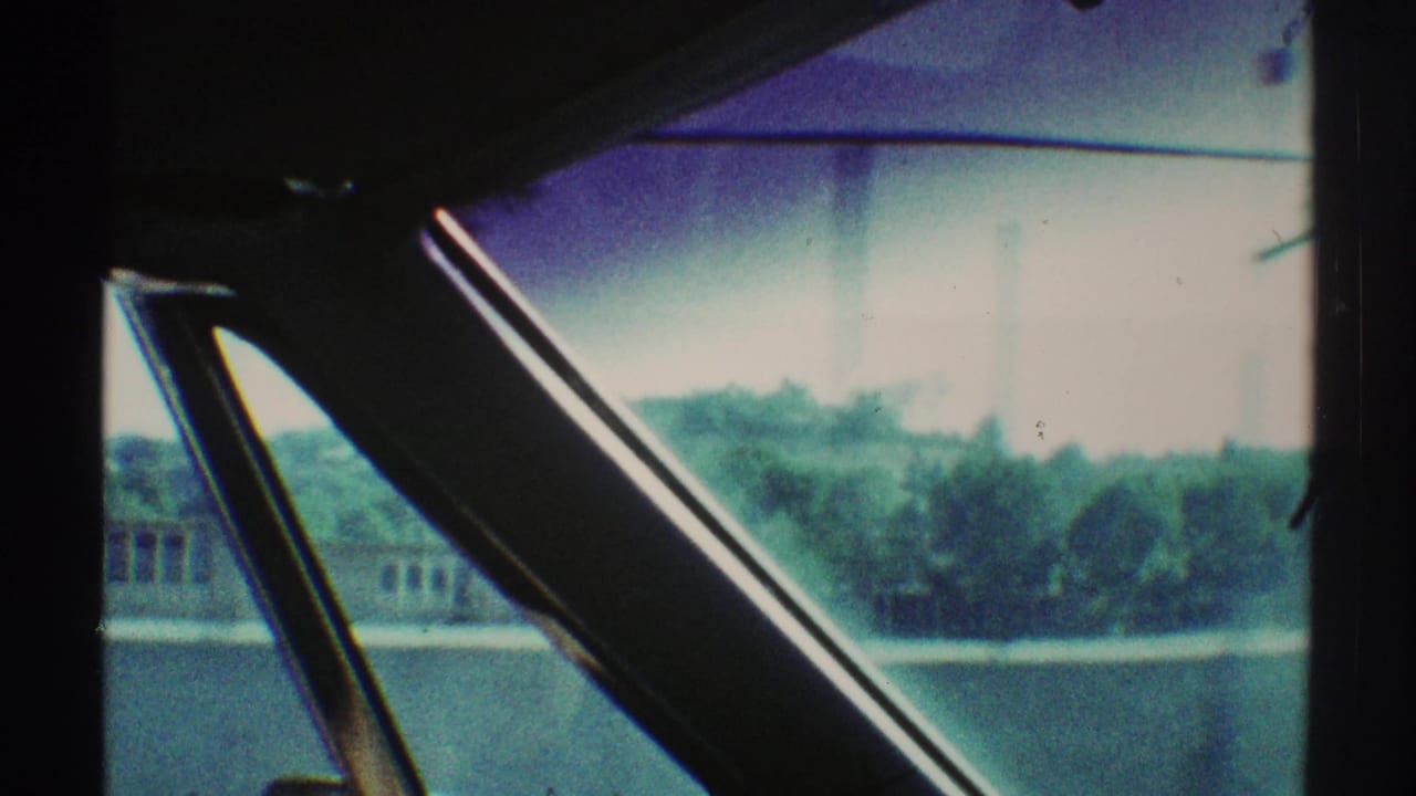 View through a car window toward trees, low buildings, and bright sky.