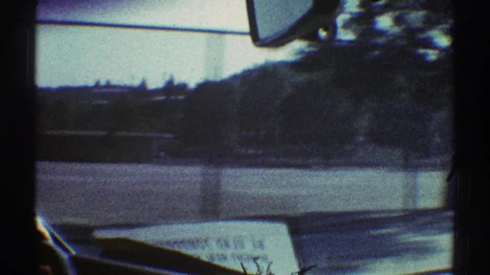 Woodland Hills driving POV in 1969, showing the windshield frame, dashboard newspaper, roadside edge, trees, and low hillside.