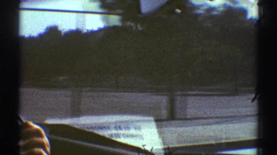 1969 car-window footage from Woodland Hills with motion-blurred trees, a dashboard newspaper, windshield frame, and a partial hand at lower left.