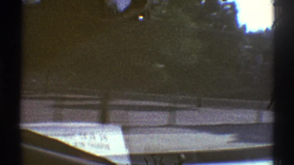 Late-1960s Woodland Hills car POV with shadowed trees outside, a dashboard newspaper inside, and the windshield frame dividing the view.