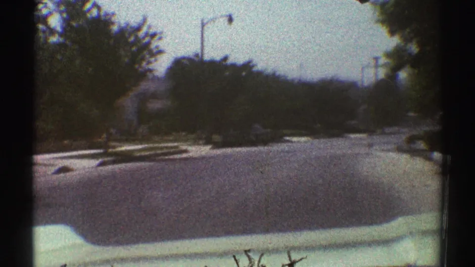 Late-1960s 8mm car POV in Woodland Hills, showing a quiet tree-lined street, lamp post, curb, and the vehicle hood edge.