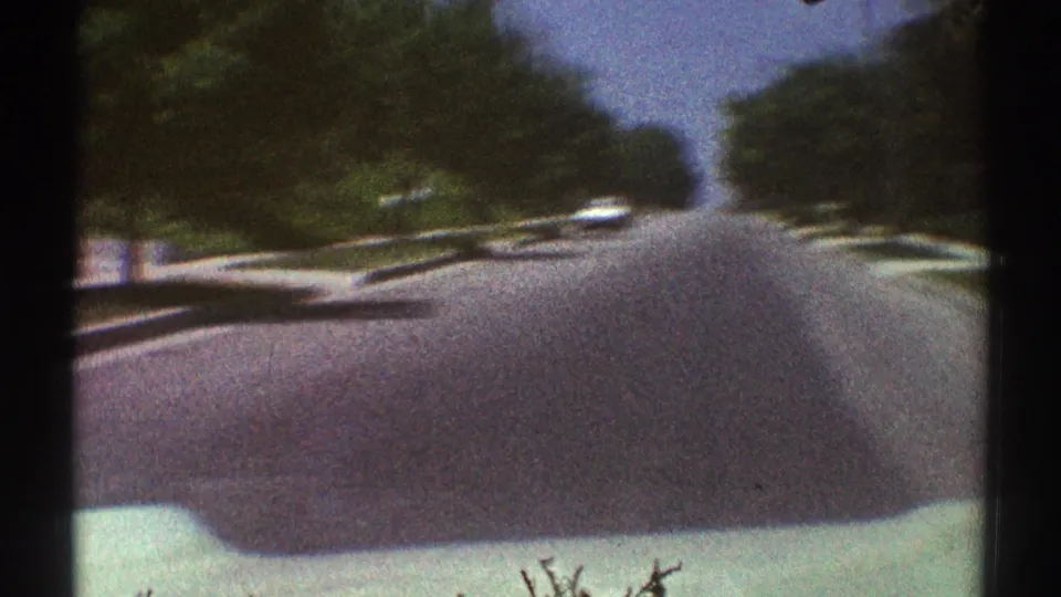 A blurred white car passes the moving camera on a shaded Woodland Hills residential street in 1969 footage.