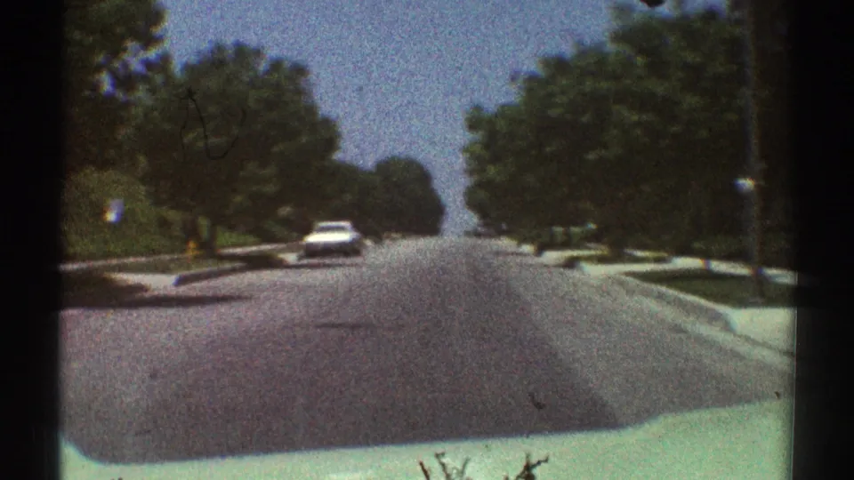 Passenger-view 8mm frame of a quiet 1969 Woodland Hills street lined with trees, sidewalks, and low residential landscaping.