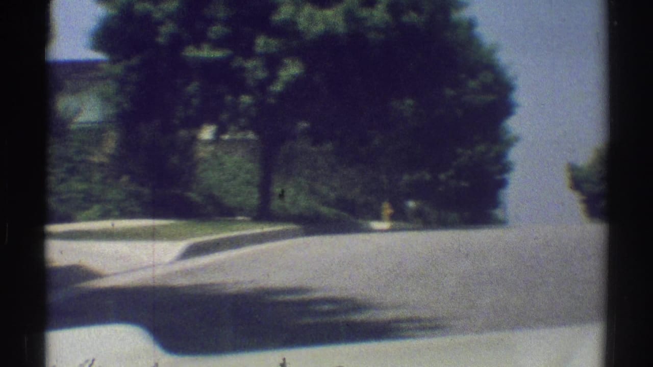 Suburban street seen from a moving vehicle with trees, sidewalk, curb, and dark film borders.