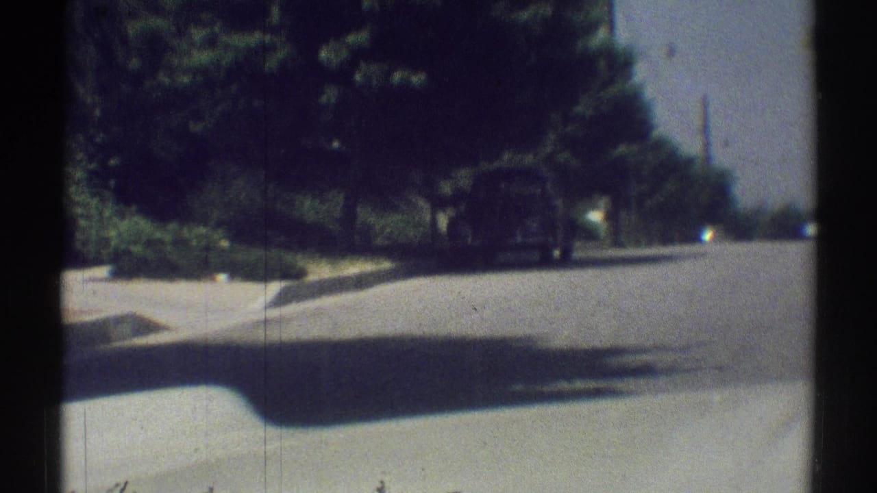 Driver’s-eye 8mm view of a quiet Woodland Hills suburban street in 1969, with trees, sidewalk, and a road stretching ahead.