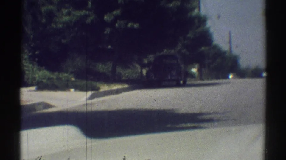 Driver’s-eye 8mm view of a quiet Woodland Hills suburban street in 1969, with trees, sidewalk, and a road stretching ahead.