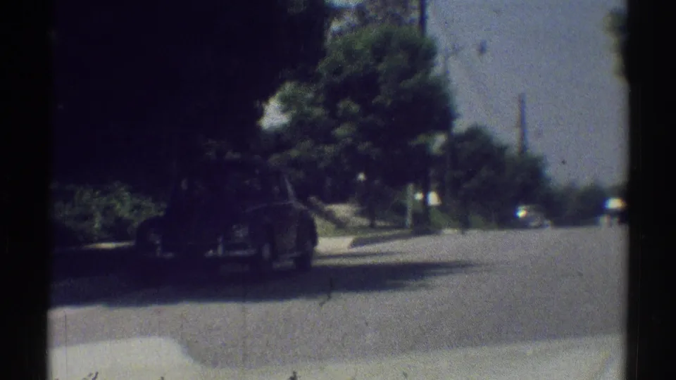 1969 Woodland Hills street view from a vehicle, with a parked car along the curb, shaded trees, sidewalk, and open road ahead.