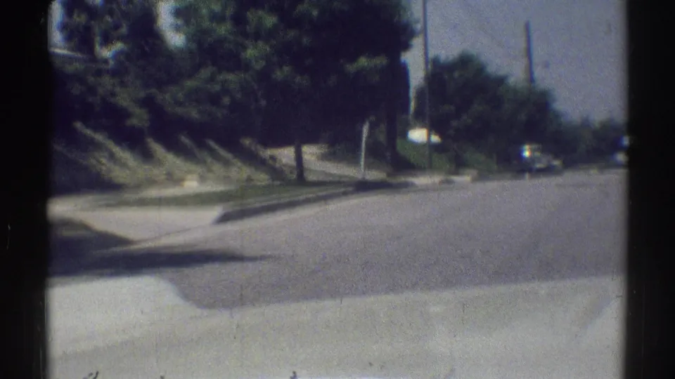 Soft 8mm driving view in Woodland Hills, showing a residential road, sidewalk, grassy hillside, trees, and a distant vehicle.