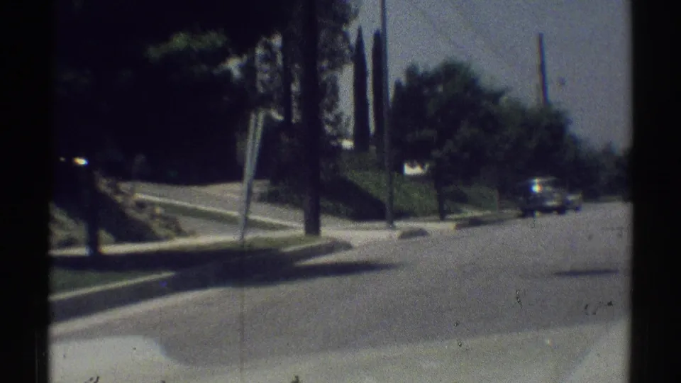 Late-1960s Woodland Hills driving frame with sidewalk, trees, utility poles, a roadside sign shape, and a car near the right edge.