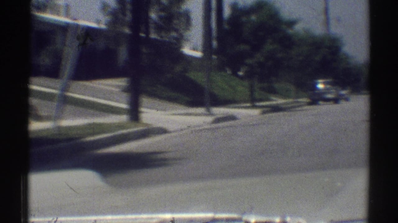 1969 Woodland Hills suburban street seen from a moving vehicle, with curbside lawns, sidewalks, trees, and a car ahead.