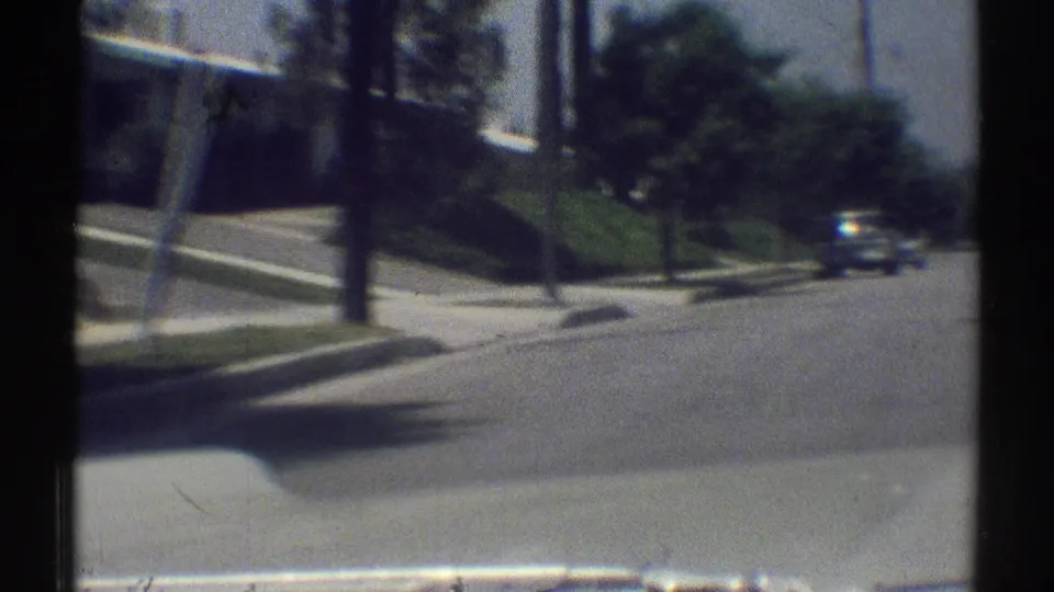 1969 Woodland Hills suburban street seen from a moving vehicle, with curbside lawns, sidewalks, trees, and a car ahead.