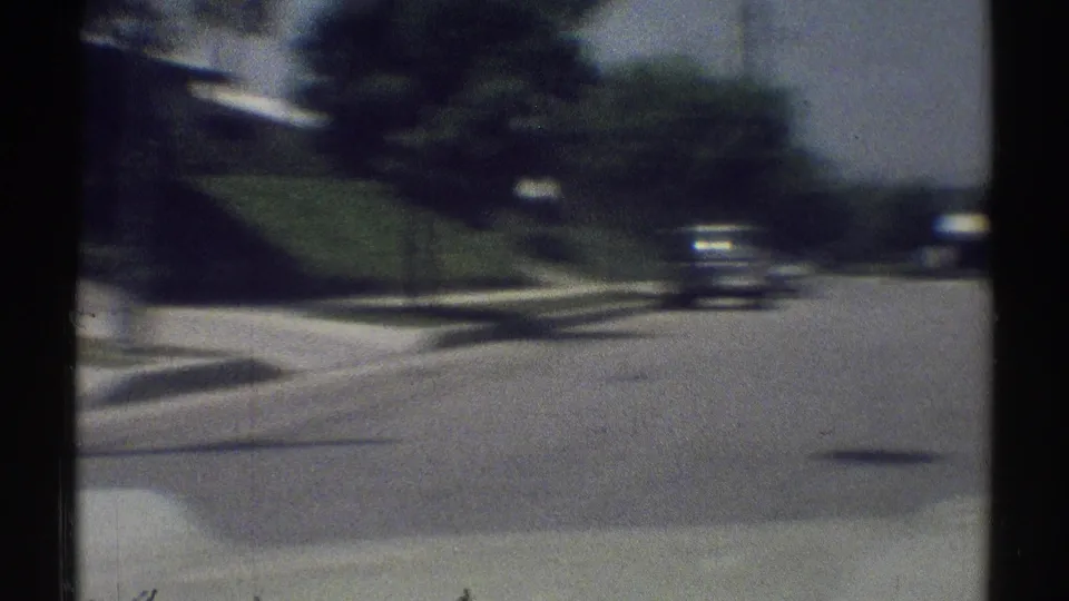 Soft 8mm view of a Woodland Hills street in 1969, showing a sloped lawn, sidewalks, trees, and a vehicle ahead.