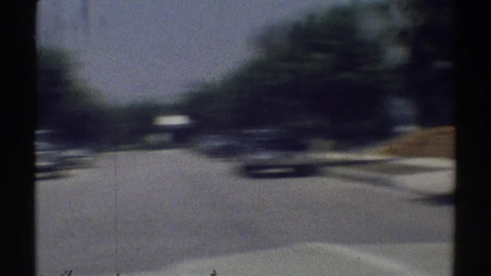 Motion-blurred 1969 suburban driving frame with passing cars, roadside trees, sidewalk, and the filming vehicle hood visible.