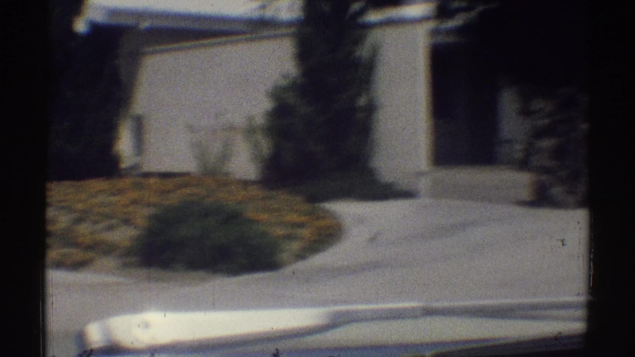 Moving car view of a white suburban house, lawn, shrubs, and walkway.