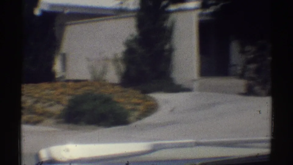1969 Woodland Hills home-movie frame showing a white suburban house, lawn, shrubs, trees, and walkway from a moving car.