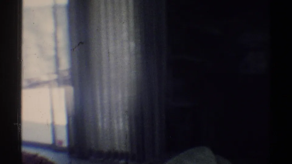 1969 Woodland Hills home-movie frame showing a dim room with a bright window, sheer curtains, and shadowed interior surfaces.