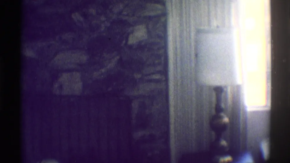 Dim 1969 Woodland Hills interior with a table lamp, bright window, curtains, and stone fireplace detail.