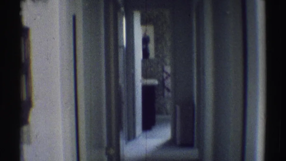 1969 Woodland Hills home-movie frame showing a dim narrow hallway with white doors, patterned wallpaper, and a person silhouetted near the far doorway.