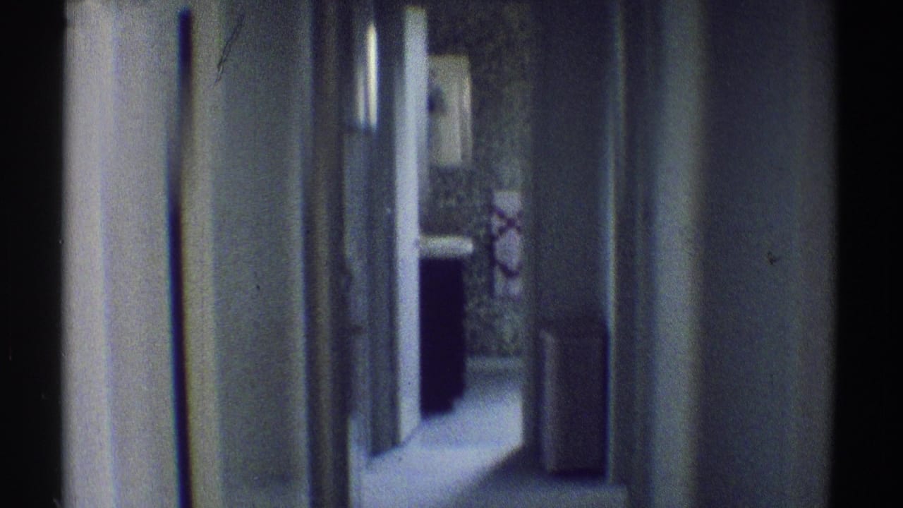 1969 Woodland Hills interior frame looking down a narrow residential hallway toward a dark-clothed person near patterned wallpaper and a doorway.