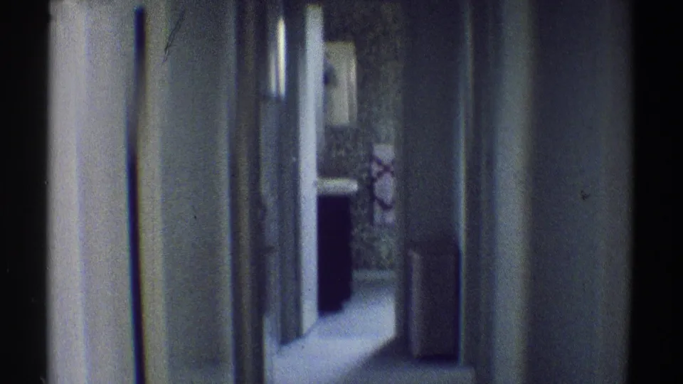 1969 Woodland Hills interior frame looking down a narrow residential hallway toward a dark-clothed person near patterned wallpaper and a doorway.