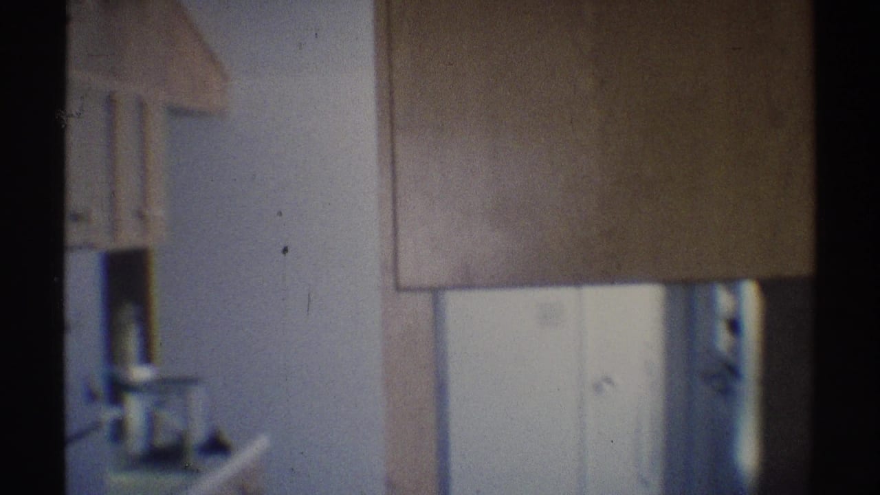 1969 Woodland Hills home-movie frame showing a dim kitchen corner with upper cabinetry, pale walls, and a refrigerator area.