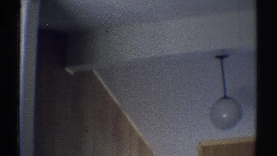 1969 home-movie frame looking upward in the kitchen toward a round pendant light, ceiling, cabinetry, and wood paneling.
