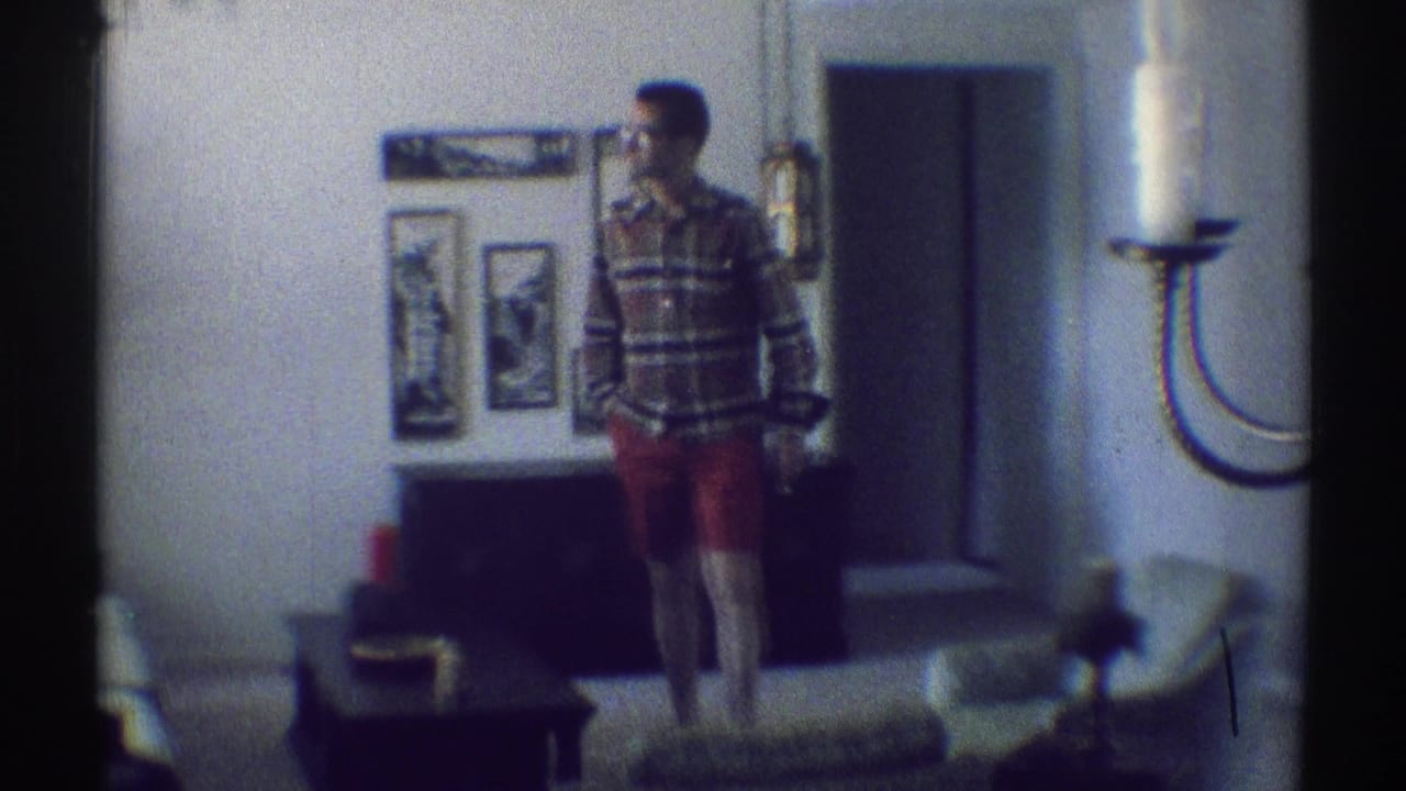 Man in plaid shirt and red shorts standing in a living room near framed wall art and a coffee table.