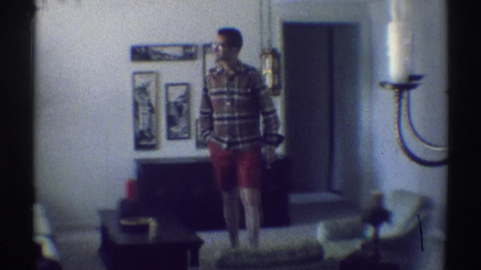 Adult man stands in a Woodland Hills living room in 1969, with framed pictures, low furniture, and a chandelier edge visible.