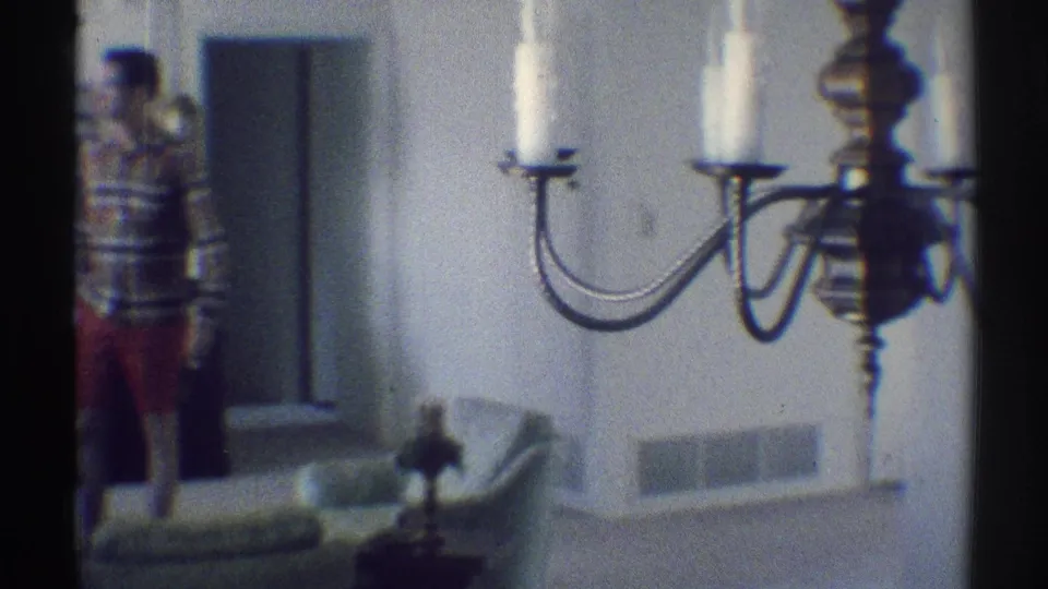 The camera angle shifts across the 1969 Woodland Hills interior, with the man at left and chandelier arms dominating the foreground.
