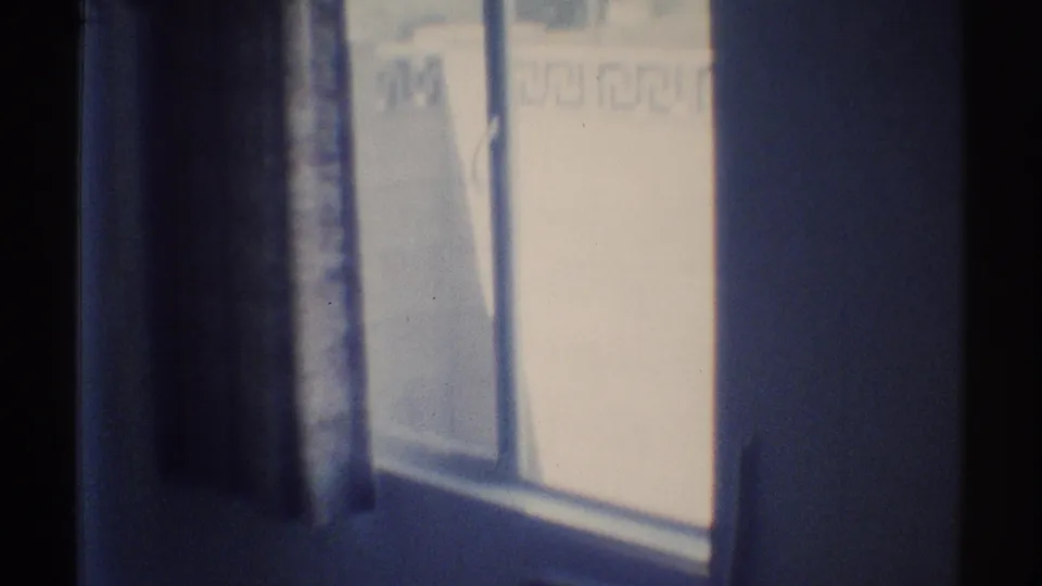 1969 Woodland Hills home-movie frame showing a dim interior wall and bright window partly covered by a dark curtain.
