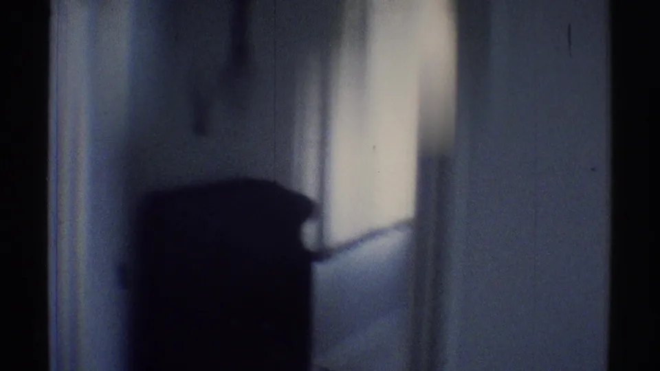 Final 8mm frame of the dim room with a dark furniture silhouette, bright window light, and shadowed wall surface.