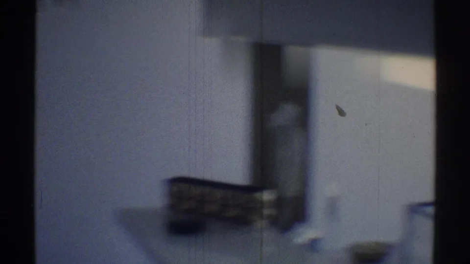 Soft 8mm frame from a Woodland Hills interior, with a wall area, dark vertical frame, and blurred household objects in the background.