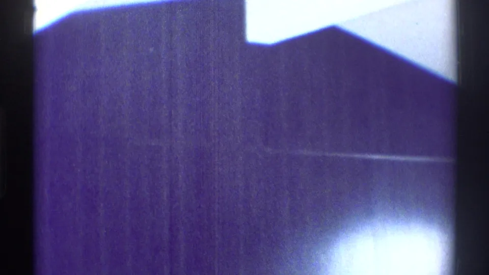 A dim 1969 interior view with a dark angular room plane, pale wall, and overexposed light bloom during the handheld pan.
