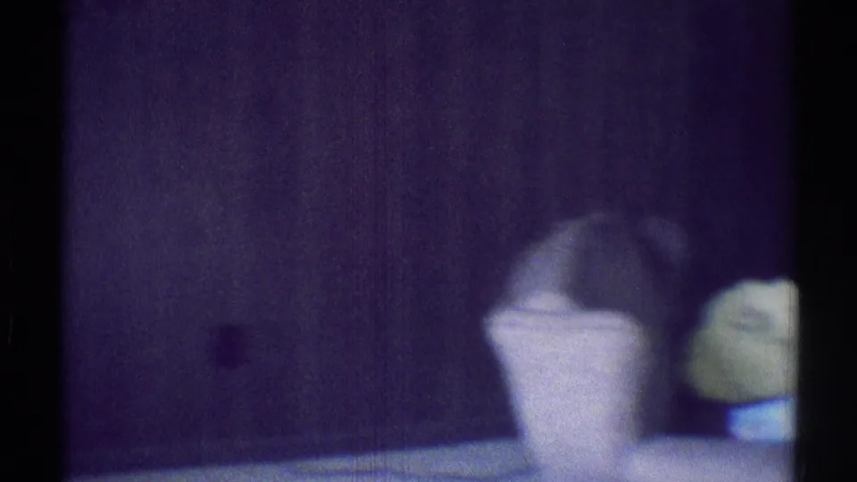 1969 8mm home-movie frame from Woodland Hills showing a dim bedroom detail with a pale pillow and green object in soft focus.