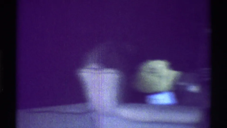Soft 8mm frame from the Woodland Hills interior, showing a pale pillow-like form and green object beside a purple-tinted wall.