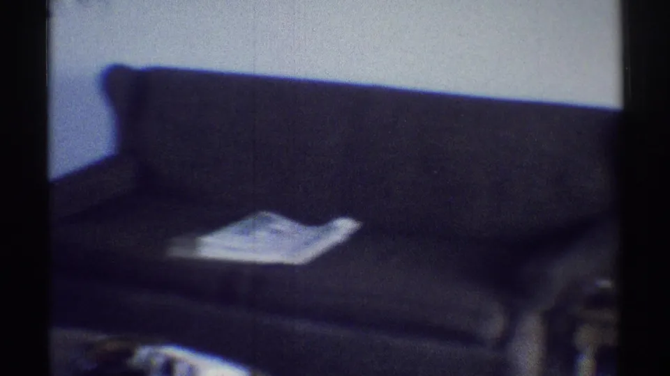 1969 Woodland Hills home-movie frame showing a dim living room with a dark sofa, carpet, and a pale paper or cloth on the seat.