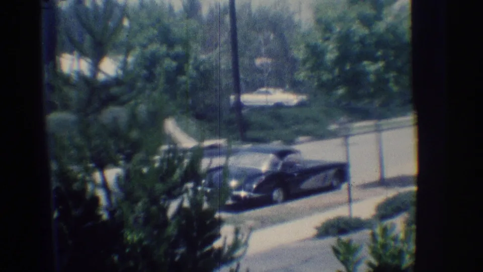 Window-framed 1969 Woodland Hills street view with a dark convertible, leafy landscaping, roadside wall, and parked cars in the distance.