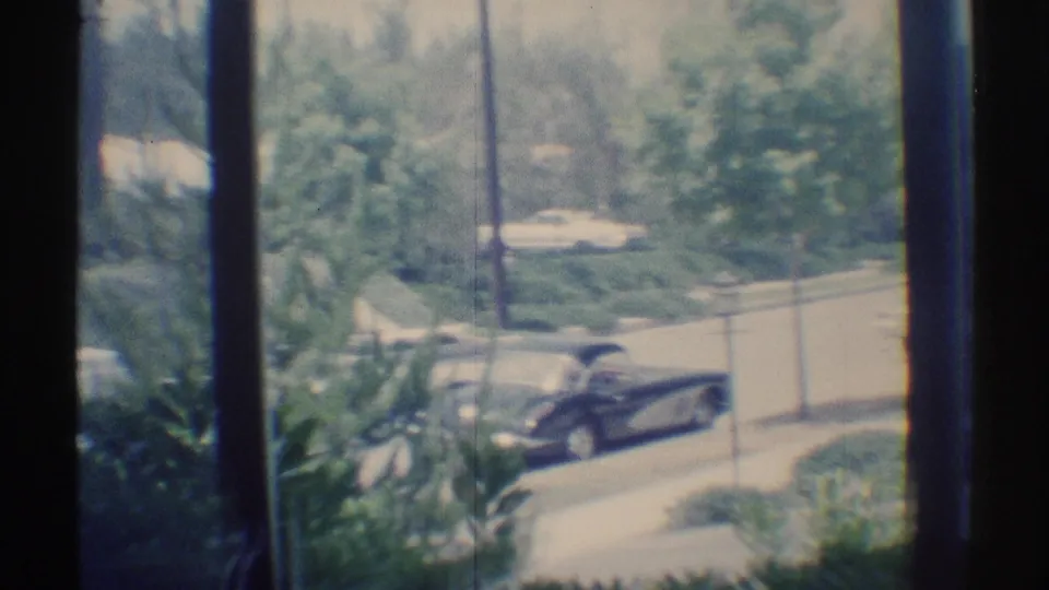 Soft 8mm frame from Woodland Hills in 1969 showing a dark car outside, shrubs in the foreground, and a residential street behind window panes.
