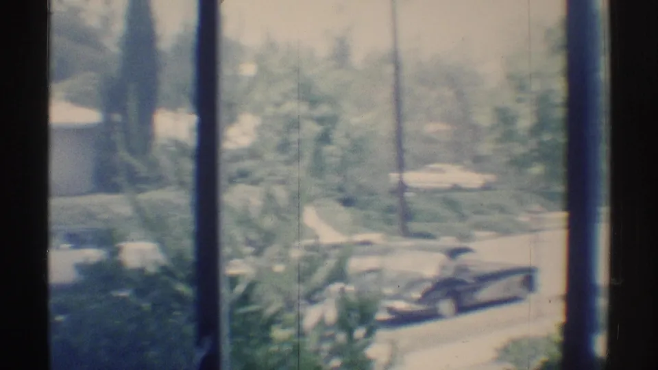 1969 home-movie frame from a Woodland Hills window, showing a dark convertible, bright wall, trees, and layered reflections or softness in the glass.